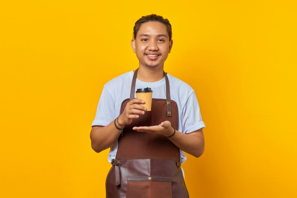 Asian barista wearing an apron holding a coffee cup against a vibrant yellow background.
