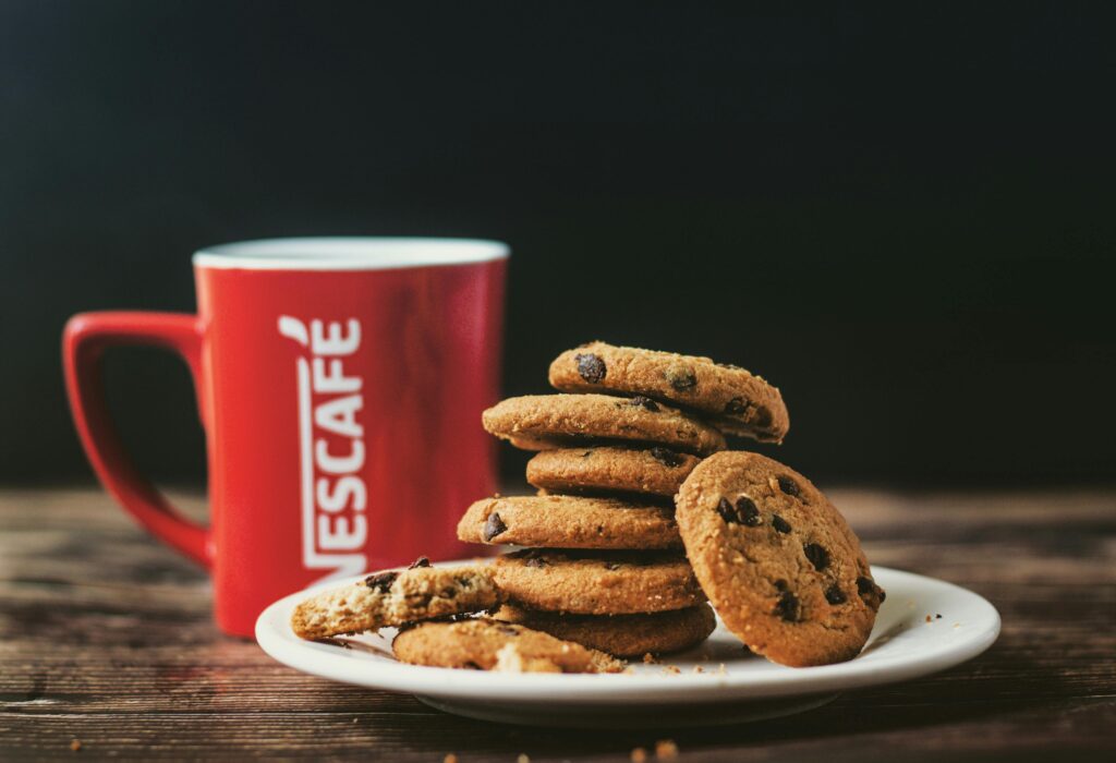 Stack of chocolate chip cookies on a plate with a red coffee cup on a dark background.