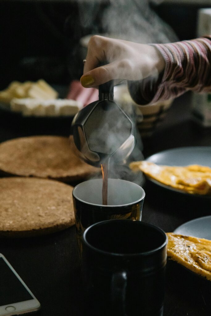 Close-up of steaming coffee being poured into a mug, surrounded by breakfast items.