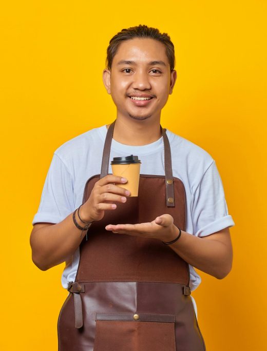 pexels-photo-11792317-11792317 Asian barista wearing an apron holding a coffee cup against a vibrant yellow background.